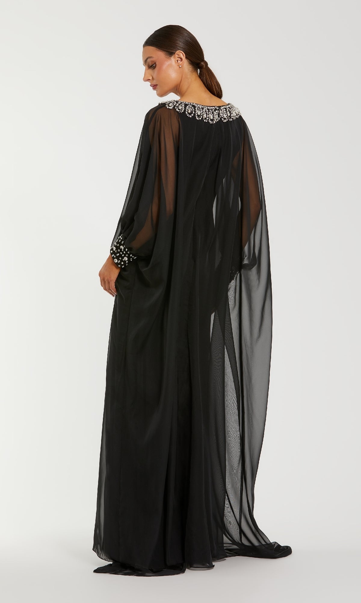 A woman wears the Mac Duggal 56203 long formal dress, featuring black chiffon fabric and rhinestone-embellished neckline and cuffs, as she stands against a white background, facing slightly away from the camera.
